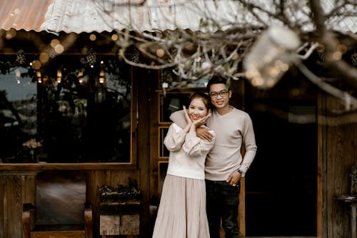Happy young husband hugging wife on terrace of wooden house near blurred leafless tree