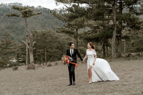 Free Full body of newlywed married Asian couple in tuxedo and wedding dress walking in green coniferous woods Stock Photo