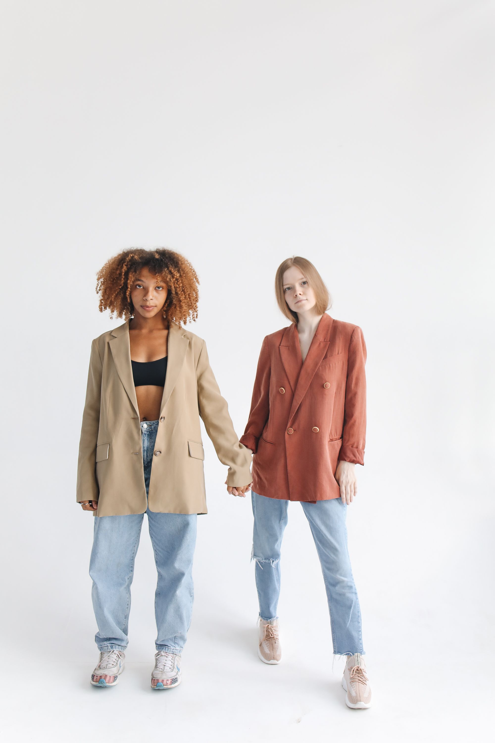 Duas mulheres estão juntas usando blazers elegantes e jeans, ilustrando um tema de amizade e moda moderna.