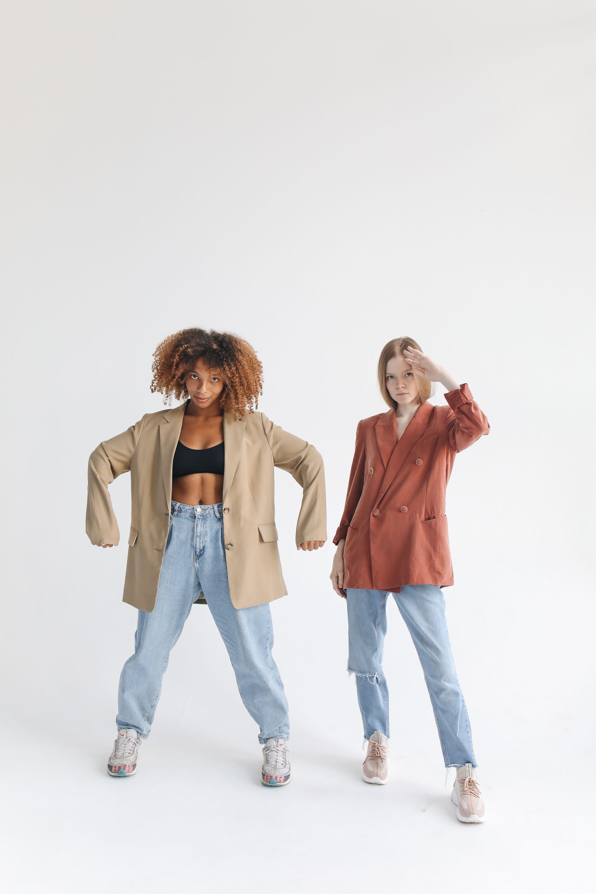 Duas modelos exibindo blazers e jeans em uma sessão de fotos de moda em estúdio, misturando estilo com confiança.
