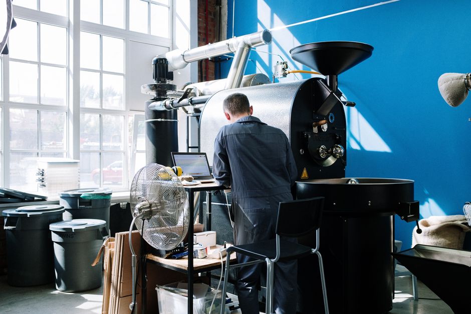 Roaster in coveralls operating a drum roaster in a sunlit workshop