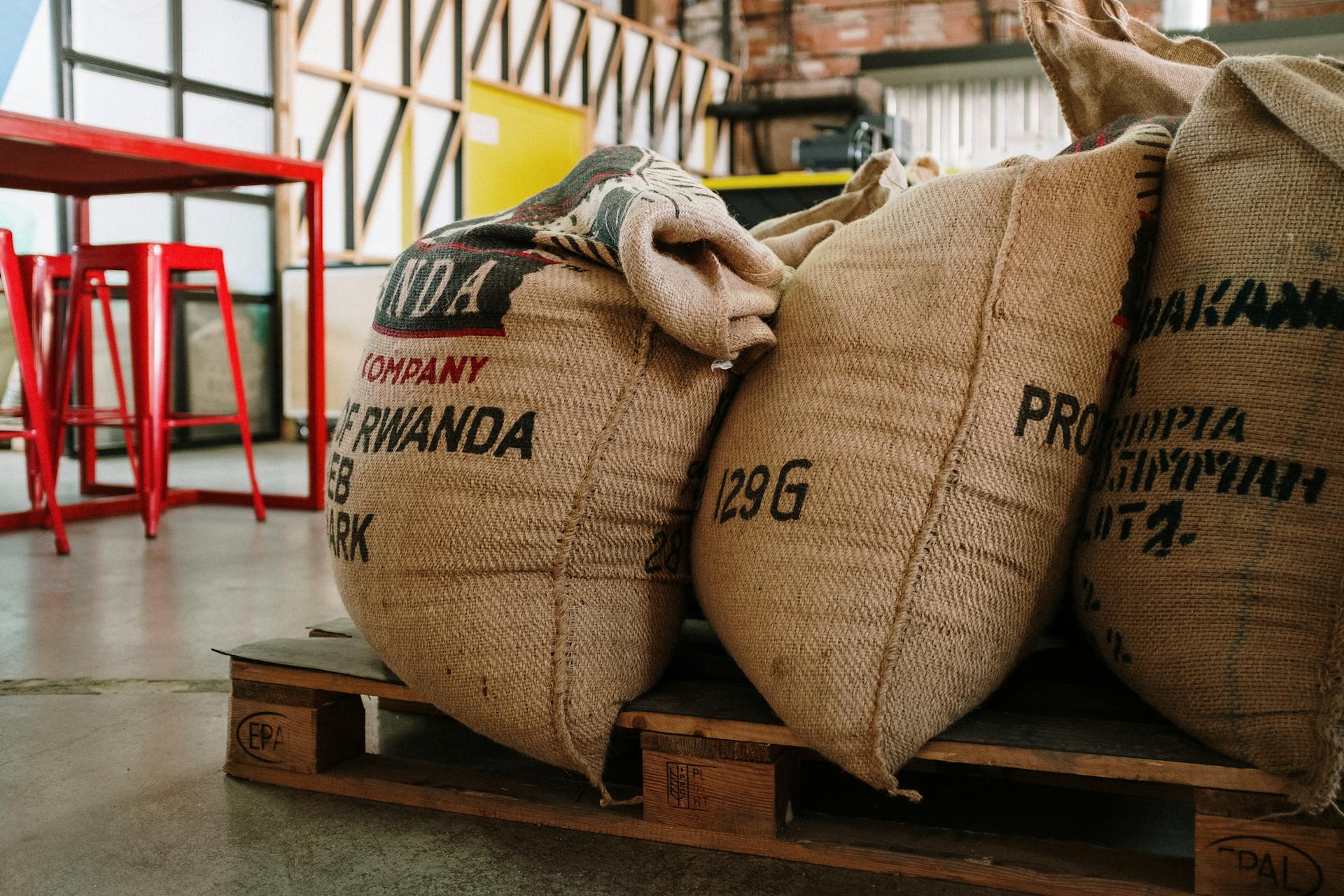 Burlap sacks of coffee beans stacked on a wooden pallet in an industrial warehouse setting.