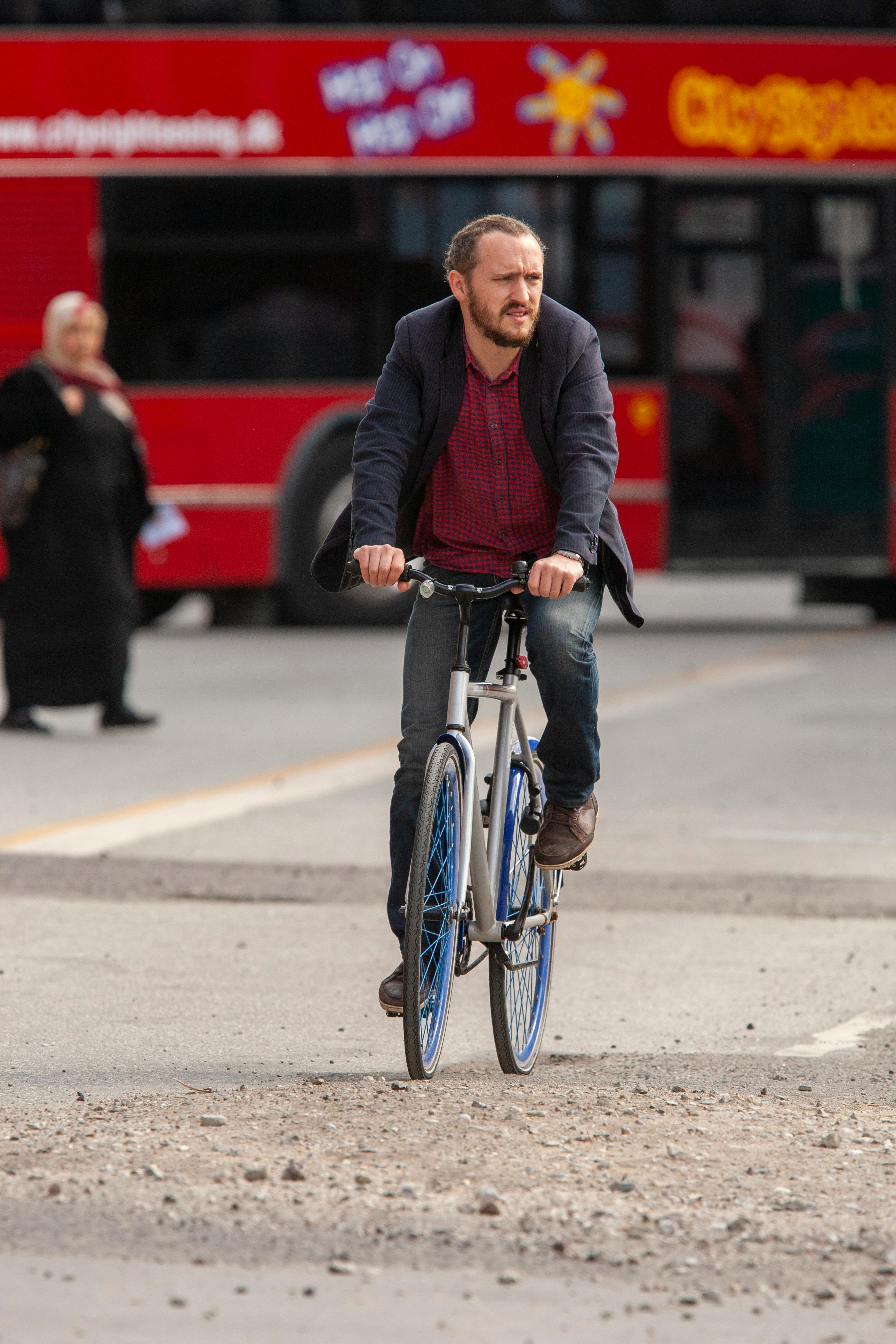 Free A man rides a bicycle down an urban street, with a red city bus in the background. Stock Photo