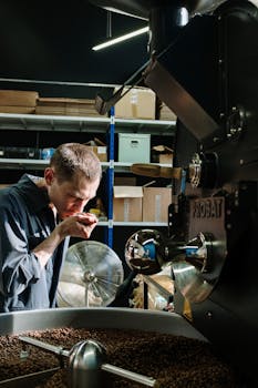 A man in a workshop smelling freshly roasted coffee beans beside industrial roasting equipment.