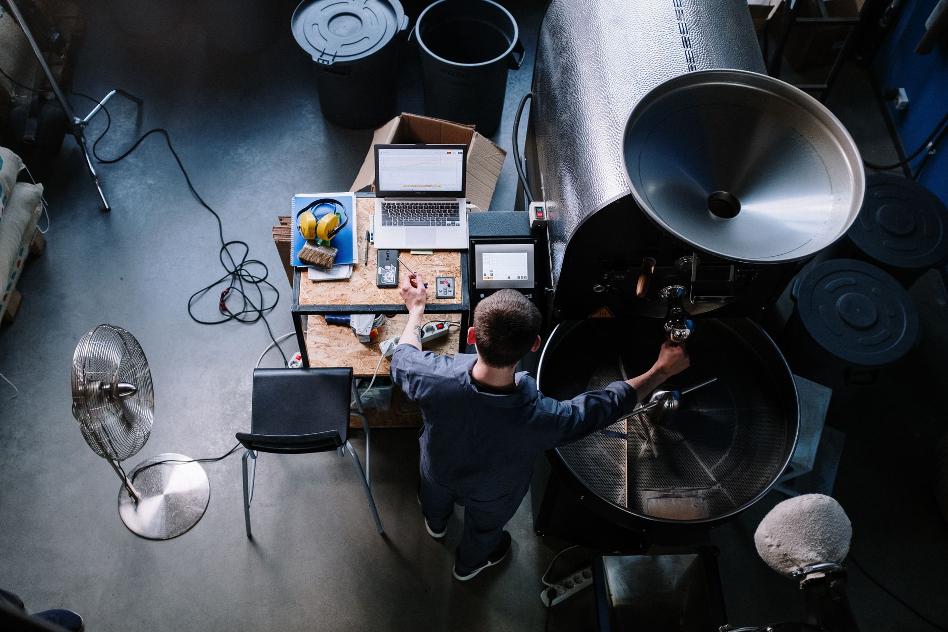 Aerial view of a man working at a coffee roaster with laptop and tools in an industrial workshop setting.