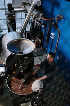 A man operates a coffee roasting machine in an industrial setting, surrounded by pipes and equipment.