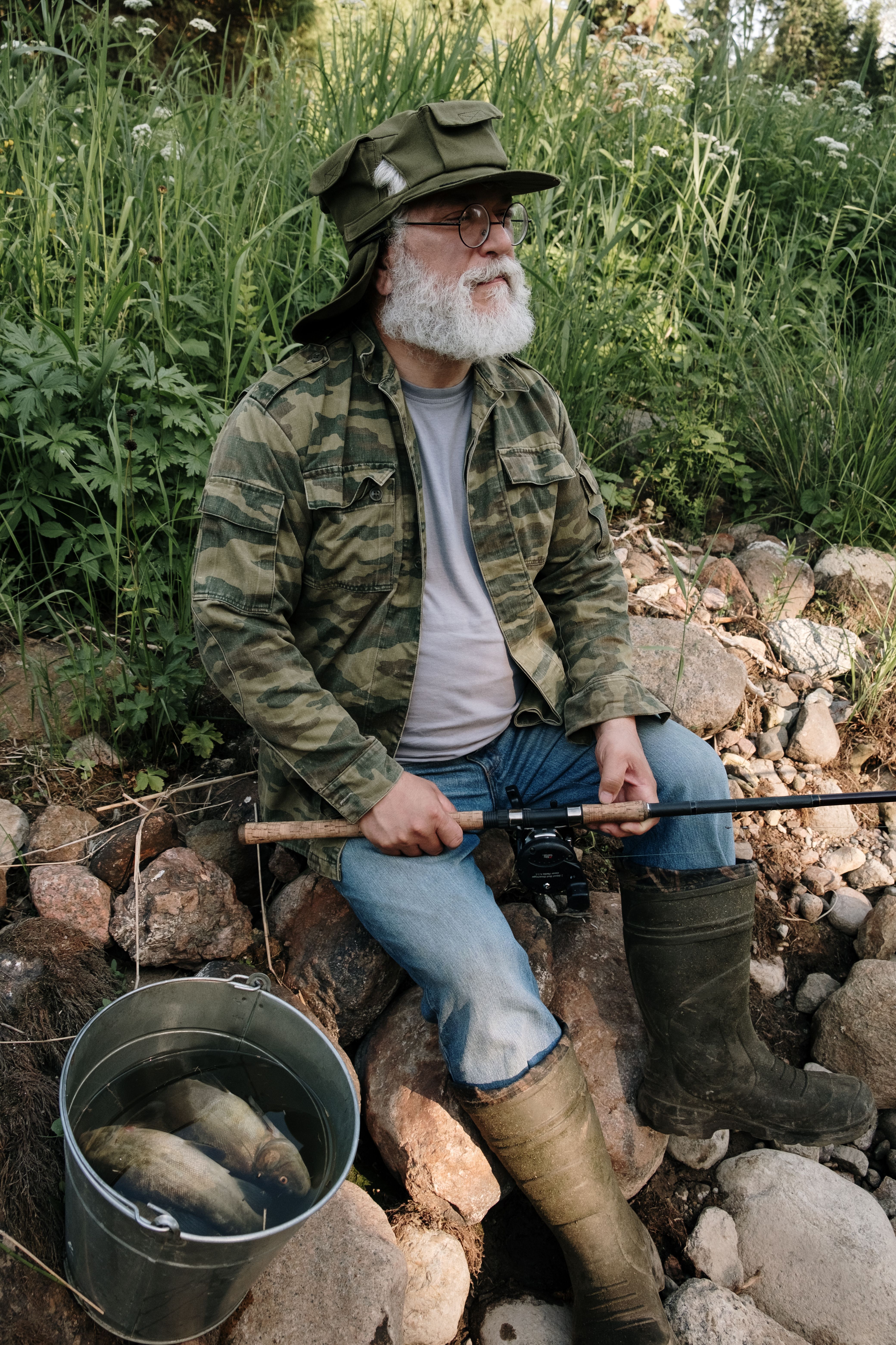 A mature man with a beard fishes at a rocky riverside wearing camouflage attire.
