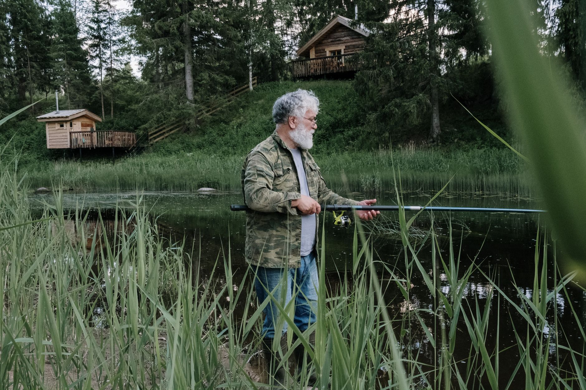 A bearded man fishing by a serene forest lake near rustic cabins.