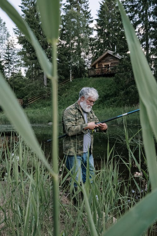 Man in Green Jacket Holding Fishing Rod Standing on Green Grass Field