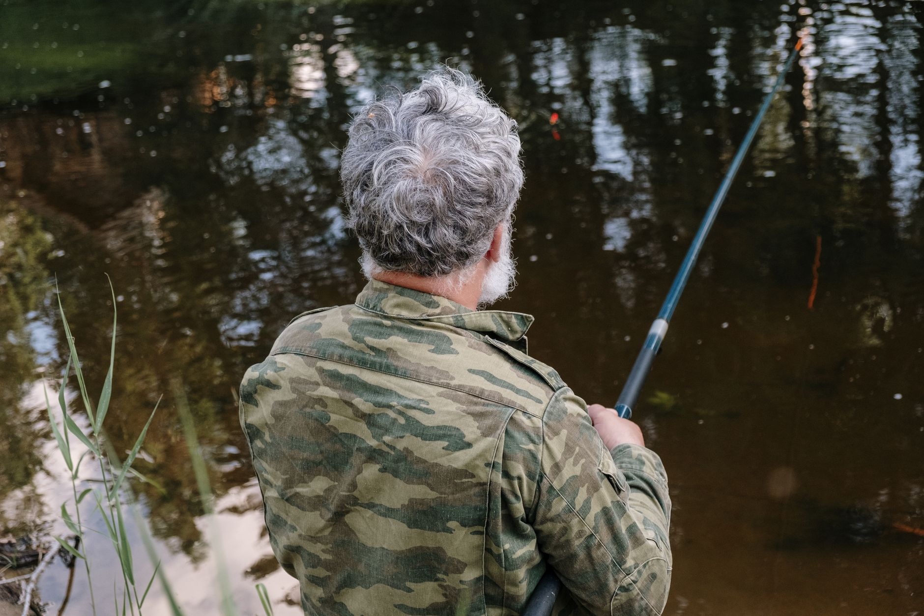 Grey-haired man in camouflage jacket enjoying a peaceful fishing session by the lake.