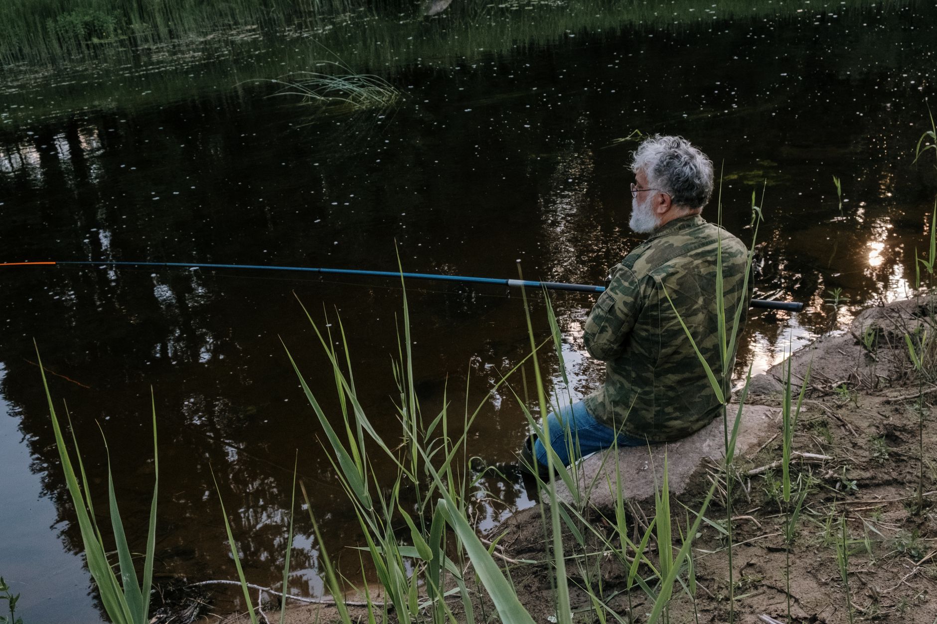 Senior man in camo jacket enjoys quiet fishing at a serene riverbank surrounded by nature.