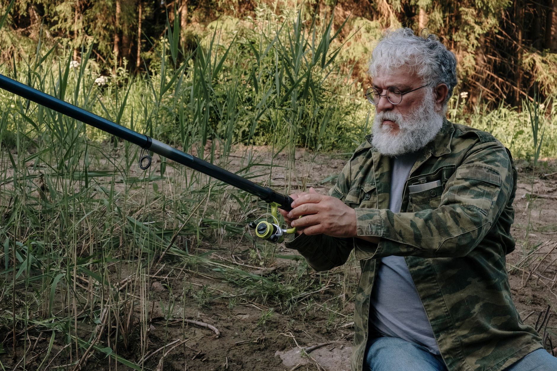 A senior man in camouflage jacket adjusting his fishing rod in natural surroundings.