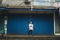 A young woman in casual attire stands in front of a blue metal shutter in Indonesia.