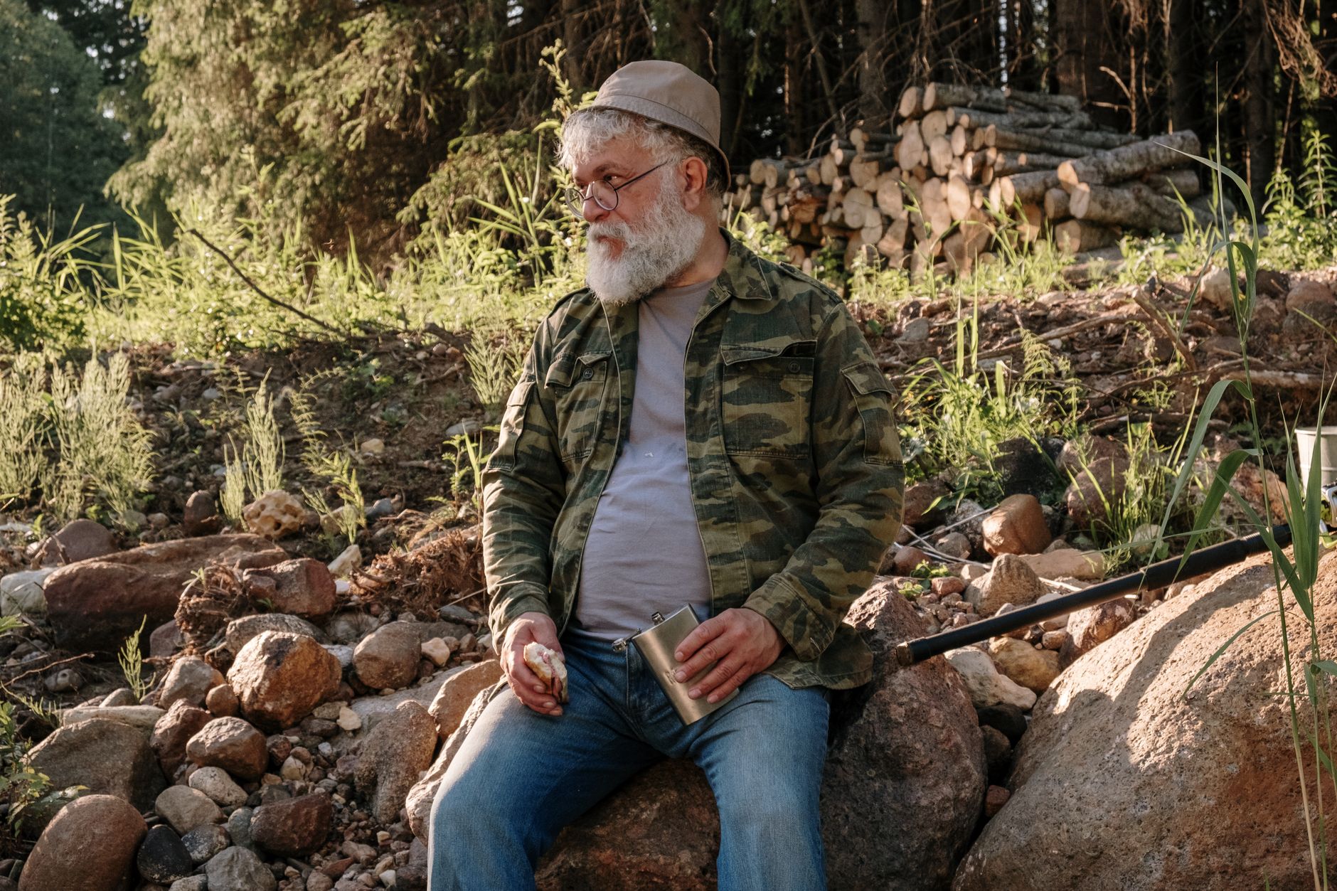 Older man sitting on rocks with a flask, enjoying nature and outdoor leisure time.