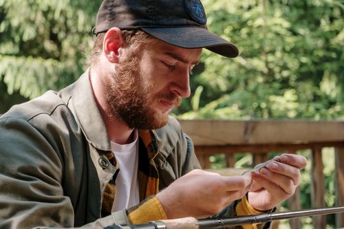 Free A bearded man focuses on preparing a fishing line outside in a serene, wooded area. Stock Photo