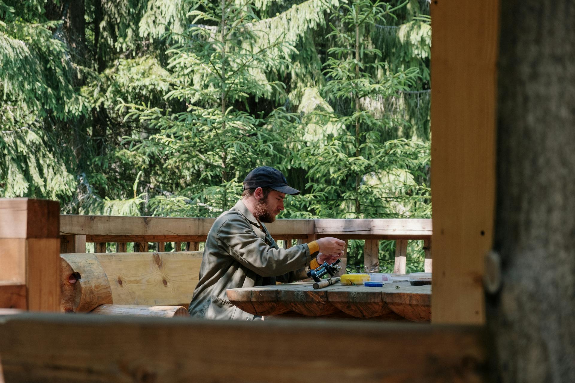 A man sits on a forest terrace handling fishing gear, surrounded by lush spruce trees.