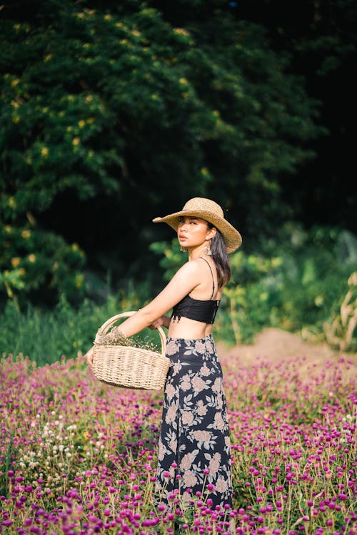 Calm female in straw hat wearing skirt and black top standing with wicker basket in hand while spending time field with flowers near green trees and looking at camera