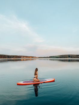 Woman in bikini paddleboarding on tranquil lake at sunset, reflecting peaceful outdoor water sport experience.