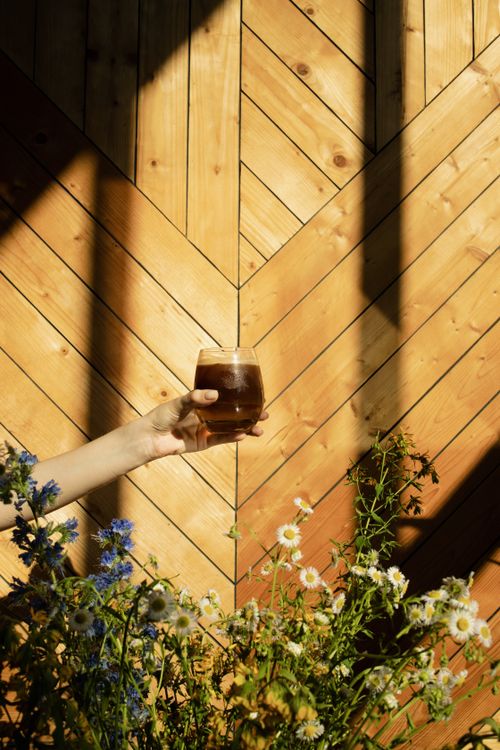 Free A hand holding a glass of iced coffee in front of a wooden wall with daisies and wildflowers. Stock Photo