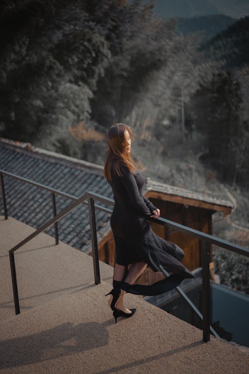 Free From above full length charming female wearing elegant black dress standing near terrace railing of rural cottage and enjoying views of lush forested hills Stock Photo