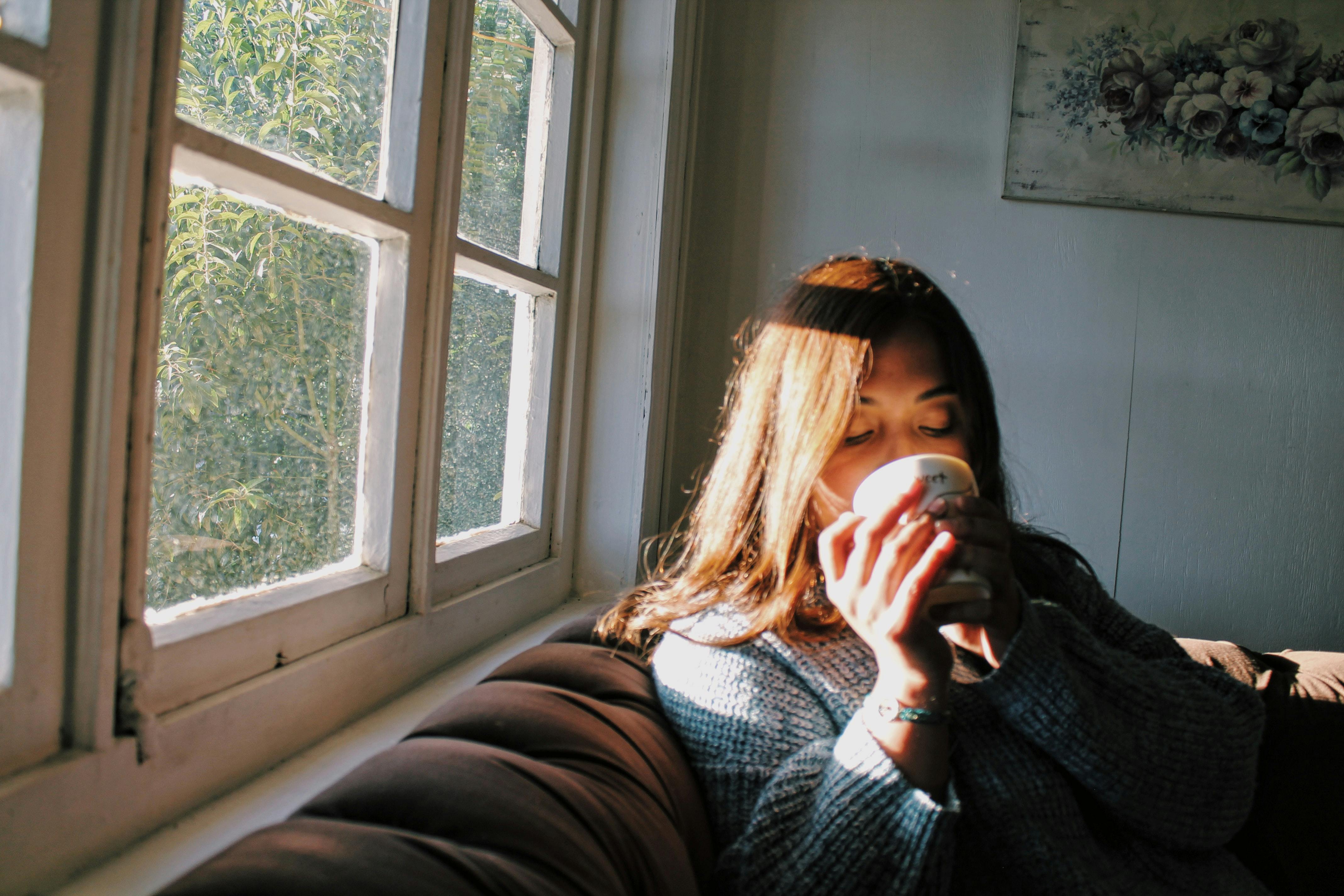 woman in cozy sweater drinking tea by window with soft natural light, calm morning aesthetic