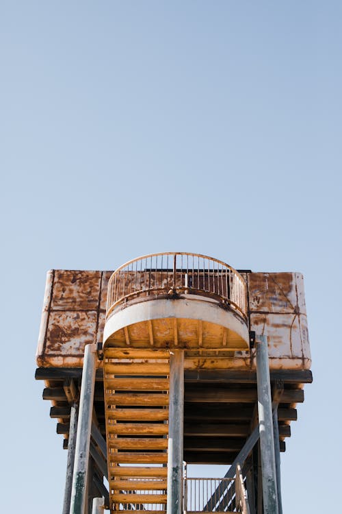 From below of aged rusty abandoned observation tower with stairway against blue cloudless sky