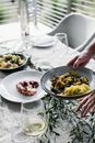 High angle of crop anonymous person putting bowl with food on table with various dishes and wineglasses and green branches