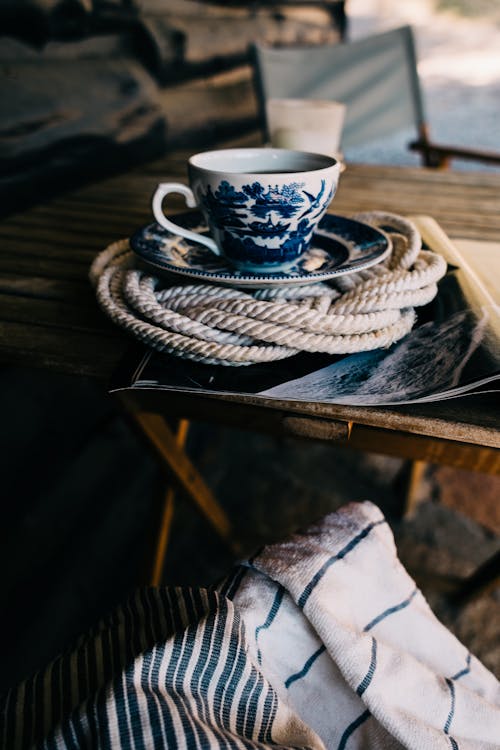 Free Ceramic cup with saucer filled with tea placed on wicker holder on wooden surface near chair in daylight Stock Photo