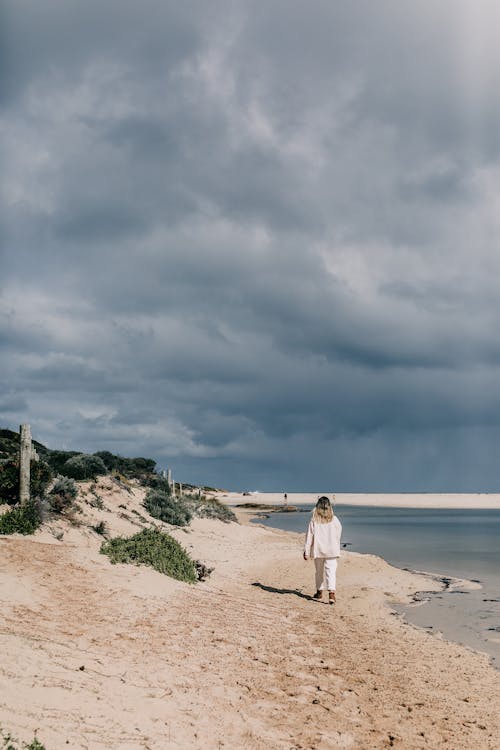 Full body back view of unrecognizable lady in white clothes walking on sandy beach near calm sea in summer day near green hills under cloudy gray sky