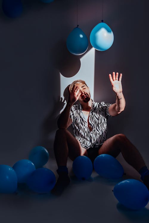 A Man Sitting on the Floor while Leaning on a Wall Surrounded by Blue Balloons