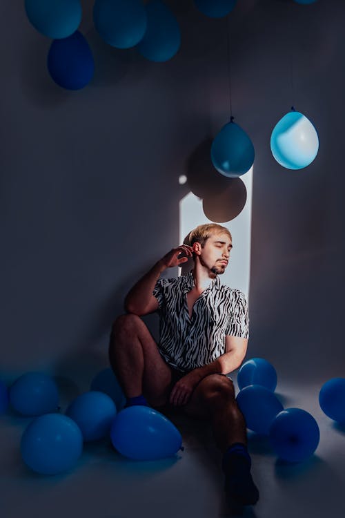 A Man Sitting on the Floor Surrounded by Blue Balloons