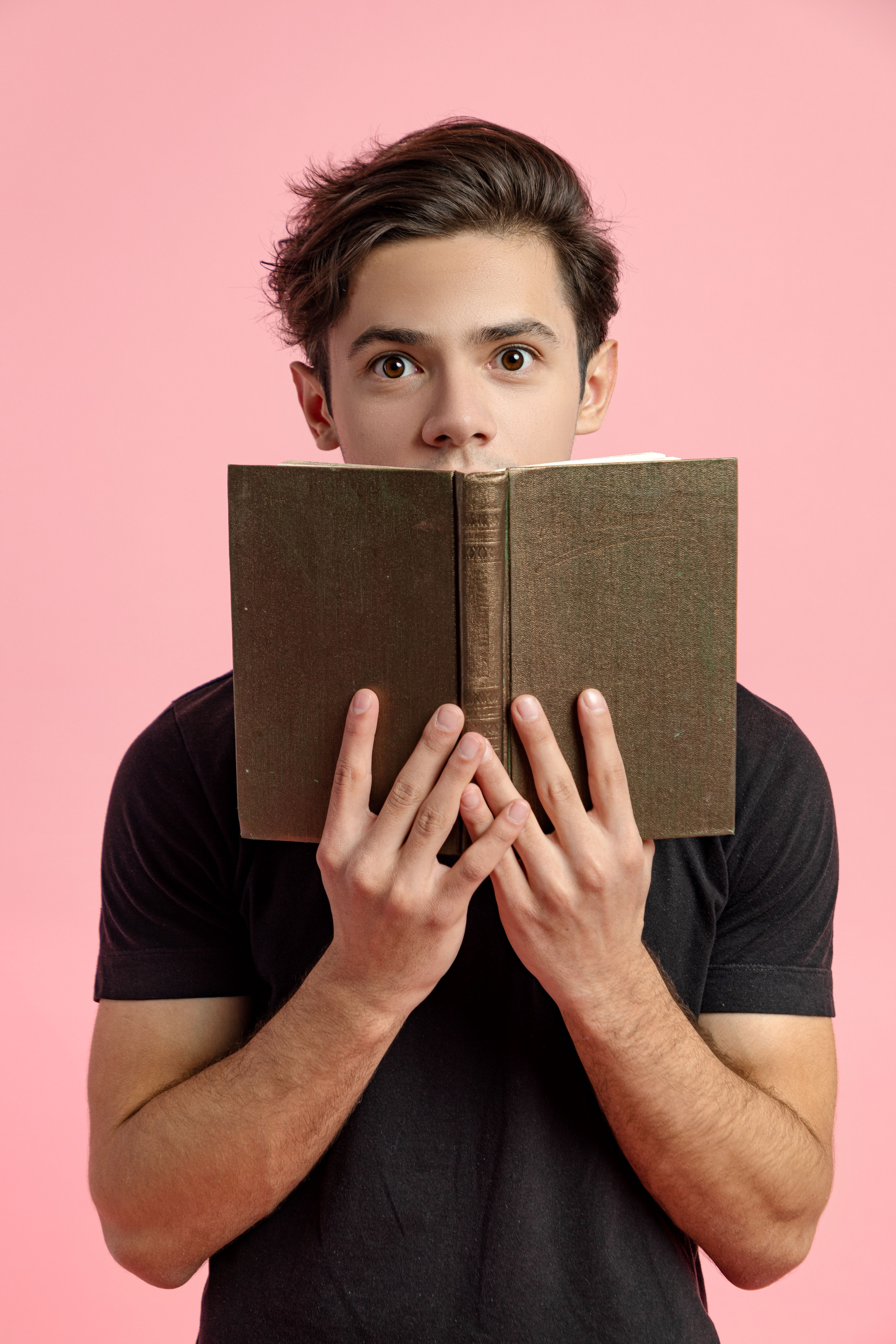 Surprised young handsome male in black shirt covering mouth with book while reading story on pink background