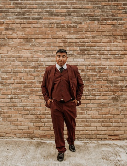 Full length confident adult male model in brown suit looking at camera while standing at wall with hands in pockets