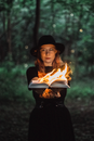 Young female wearing black clothes standing with burning pages of opened book in woodland