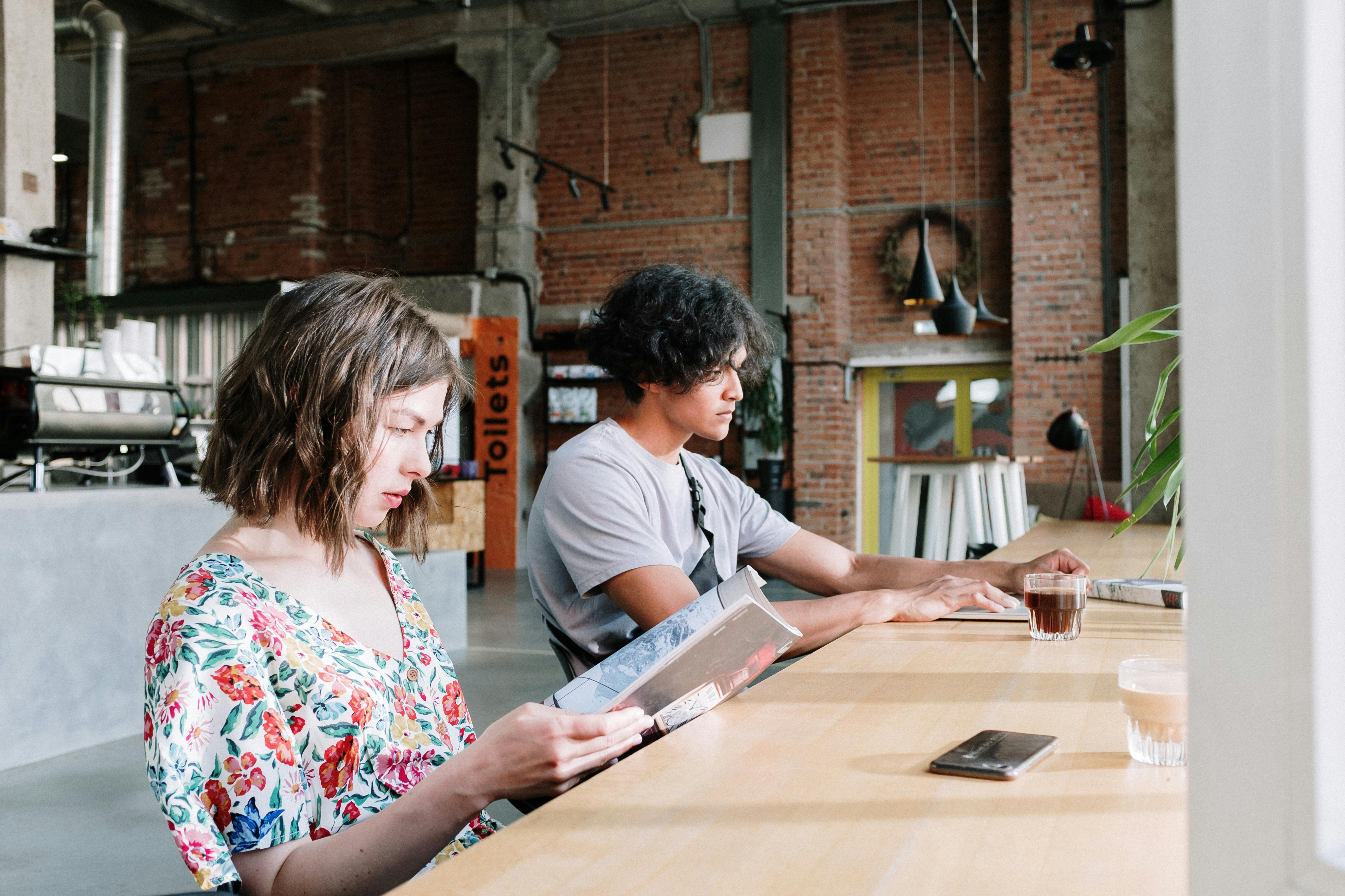A man and woman enjoy reading and coffee in a stylish industrial café setting.