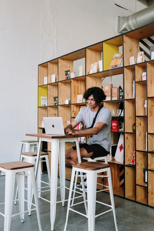 Free A young man focused on his laptop while sitting in a stylish, modern café with wooden shelving. Stock Photo