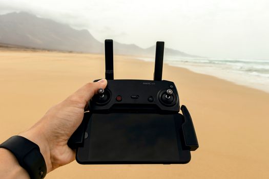 A hand holding a drone controller with a scenic sandy beach background.