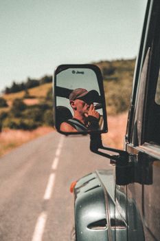Reflection of a man smoking in a Jeep mirror driving on a scenic rural road, summer day