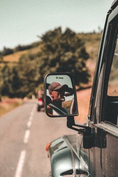 Close-up of a man smoking while driving a jeep, captured in the side mirror on a scenic road.