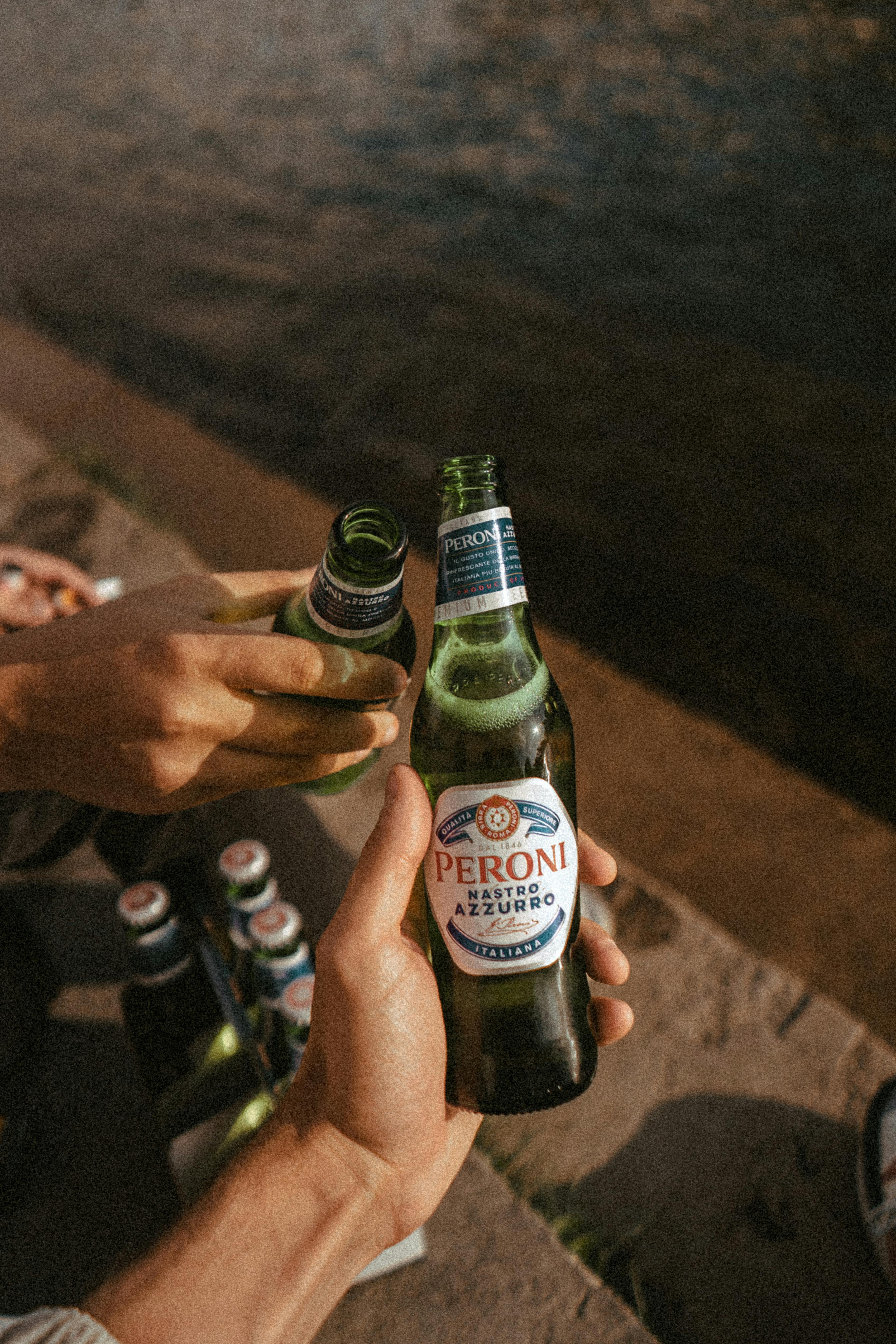 Two people clink Peroni beer bottles by the waterside, enjoying a sunny day outdoors.