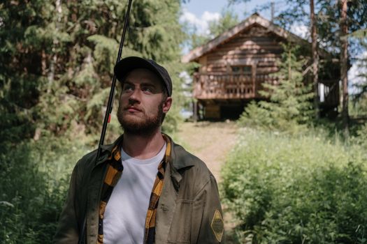 Bearded man holding fishing rod in lush greenery by a log cabin.