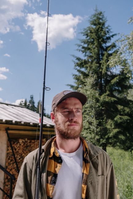 Bearded man holding a fishing rod in a rural setting with trees and a wooden cabin on a sunny day.