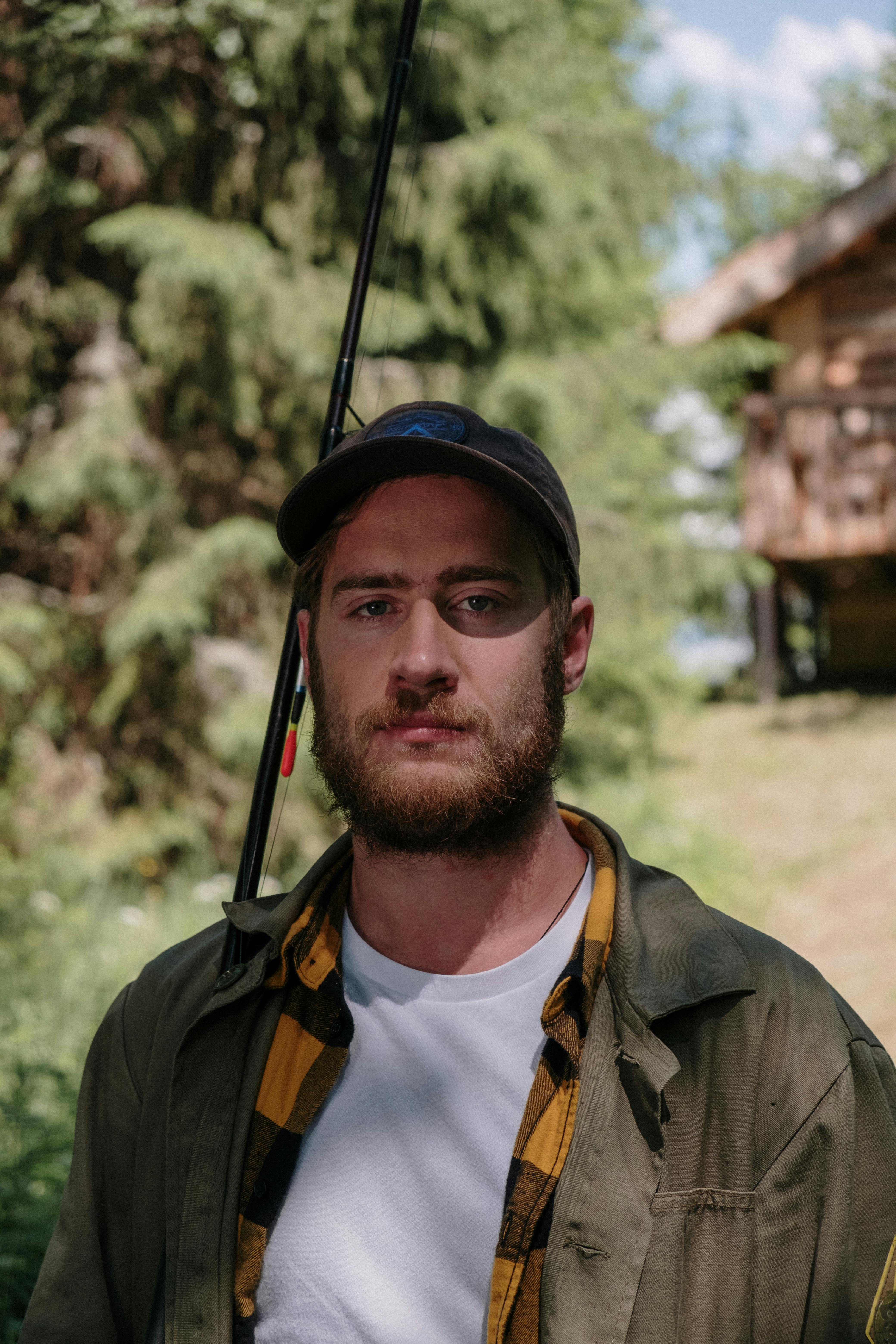 Portrait of a bearded man outdoors with fishing gear, showcasing a rustic countryside setting.