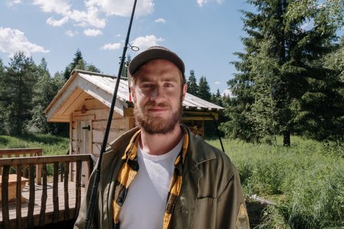 Free A bearded man holding a fishing rod outside a wooden cabin in a green summer countryside. Stock Photo
