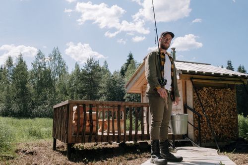 Free A bearded man with a fishing rod stands by a wooden cabin in nature. Stock Photo
