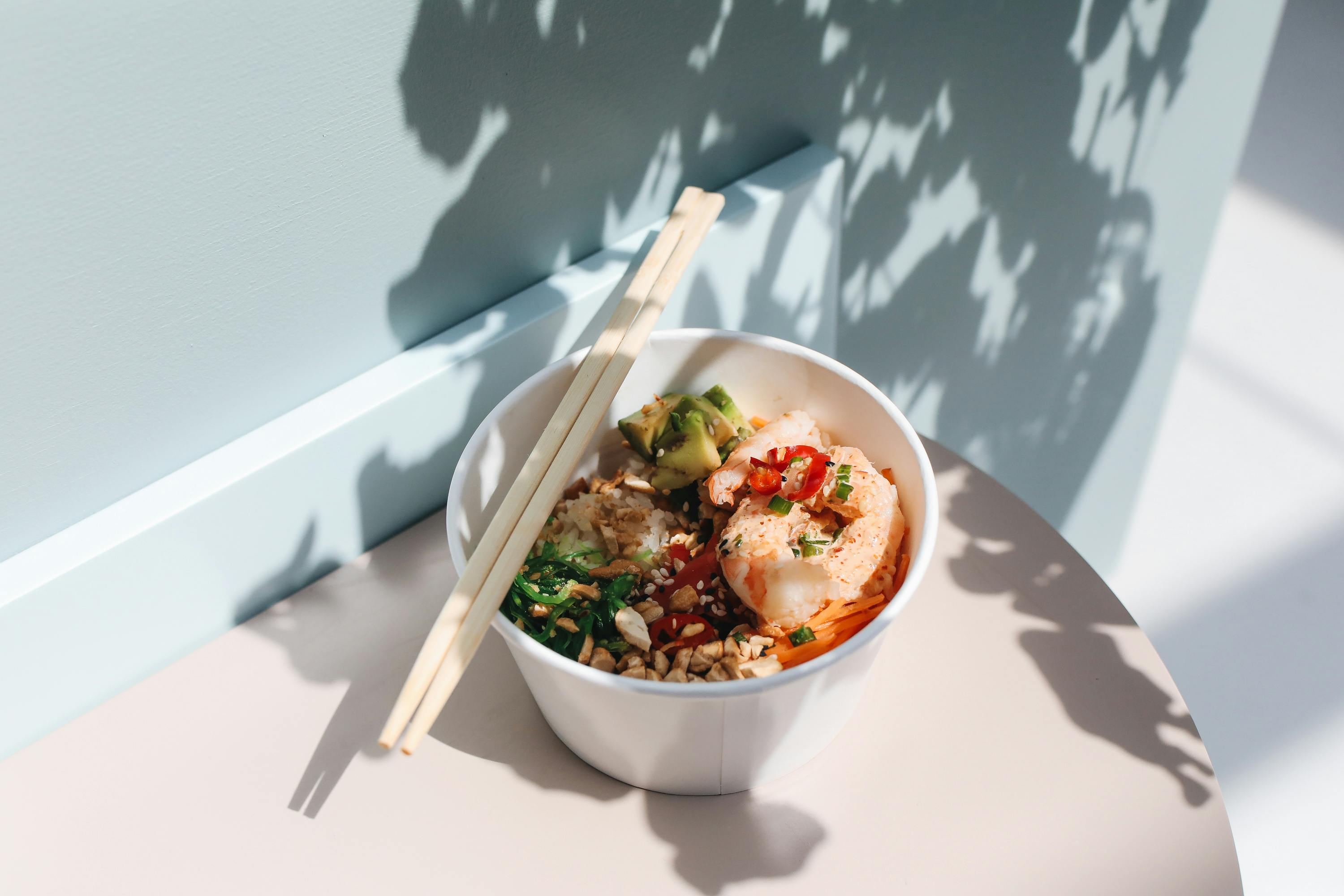 Delicious poke bowl with shrimp, avocado, veggies, and rice, served with chopsticks on a sunny day.