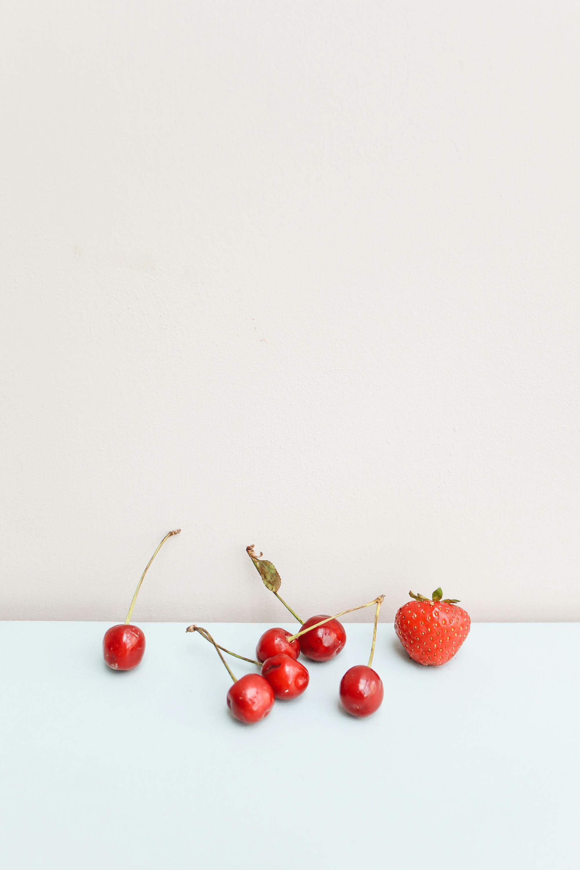 A collection of fresh red cherries and a strawberry on a minimalist pastel background.
