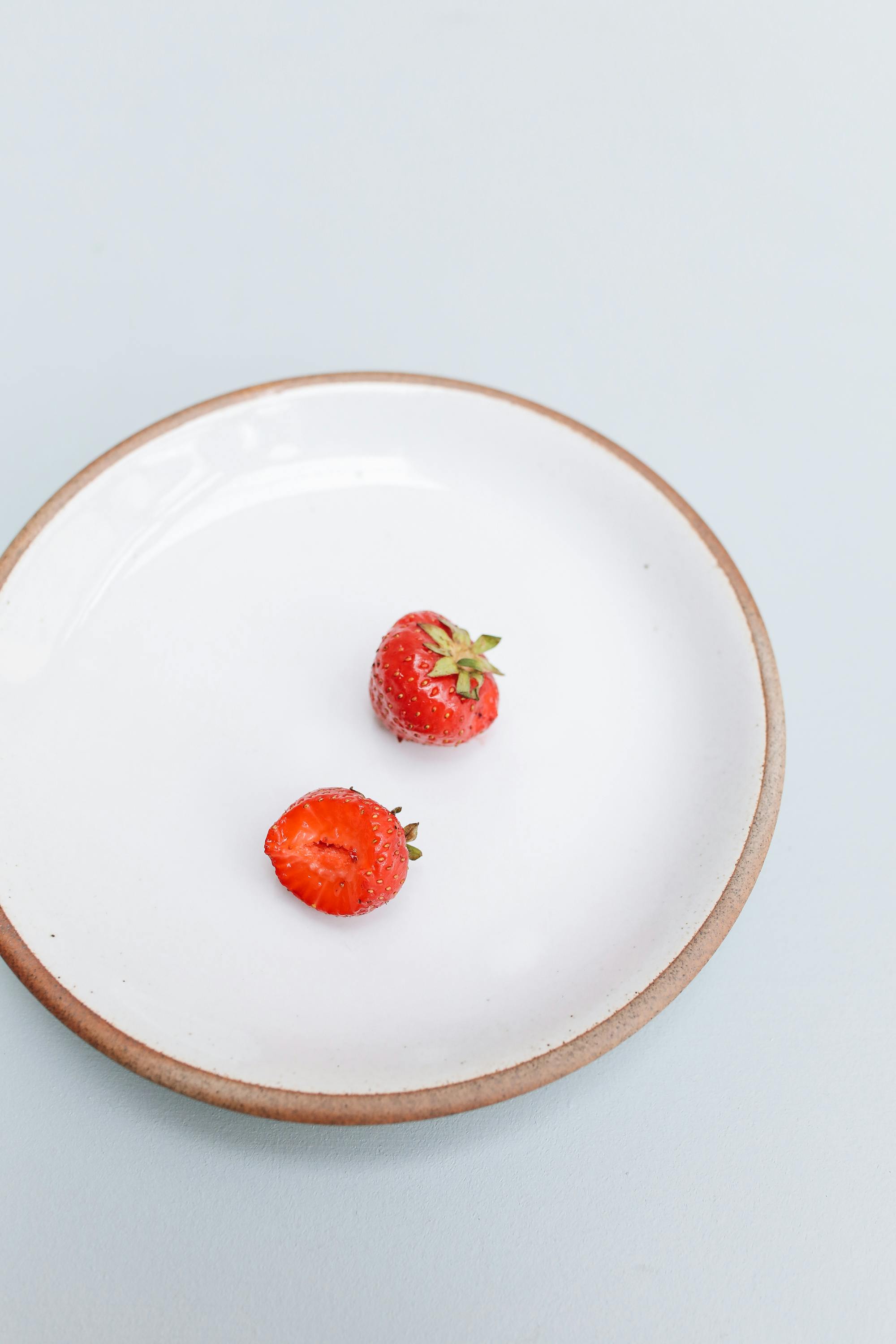Two strawberries, one bitten, on a white ceramic plate against a pastel background.