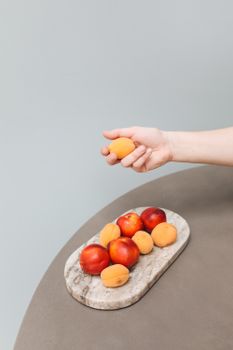 A hand delicately holds an apricot above a marble tray with assorted fruits, offering a fresh and minimal aesthetic.