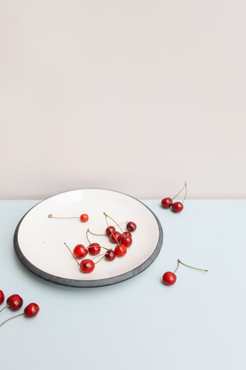 Free Fresh cherries scattered on a white plate over a pastel surface, showcasing minimalist food styling. Stock Photo
